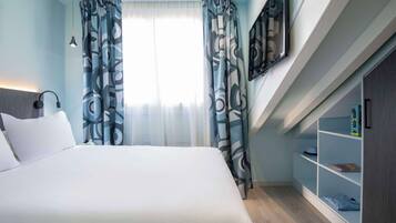 A modern hotel room with a large bed, a desk, a bookshelf, and a window with patterned curtains.