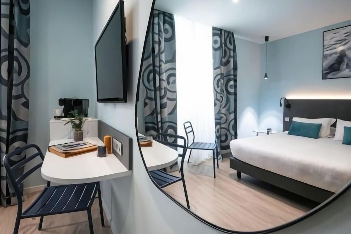 A modern bedroom with a bed, a desk, and a chair. There is a TV mounted on the wall and a window with curtains.