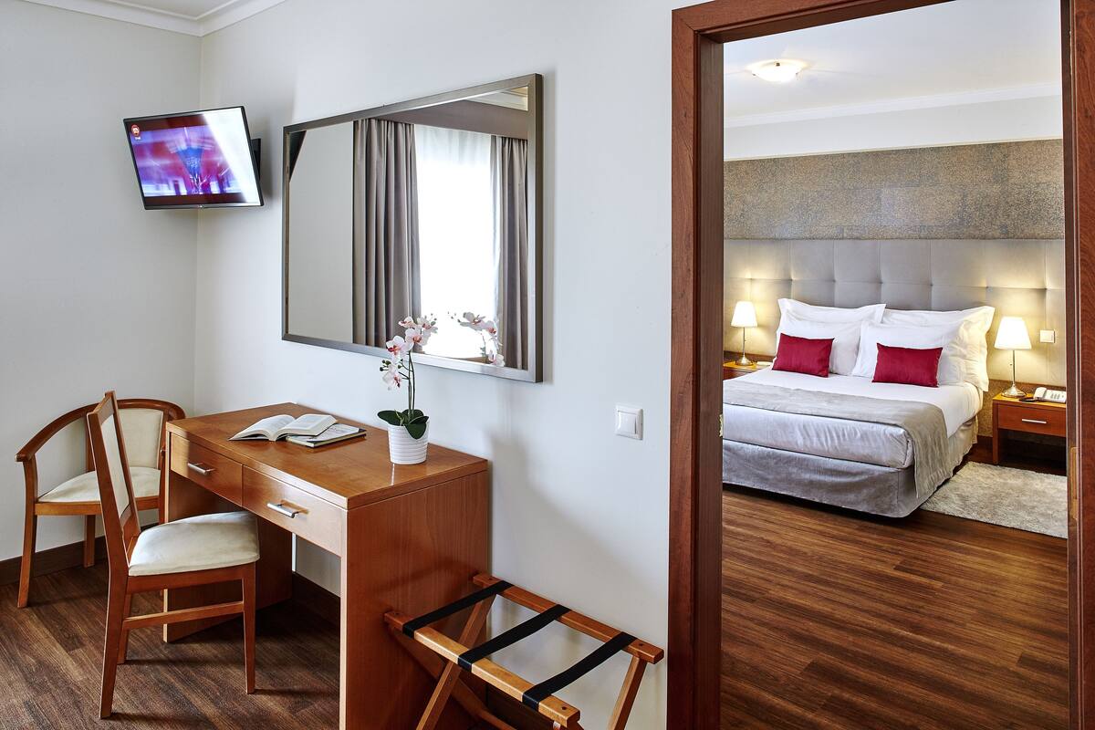 Una camera d'albergo con una scrivania in legno, due sedie, un letto con cuscini bianchi e rossi, un comodino con una lampada e un televisore fissato alla parete.