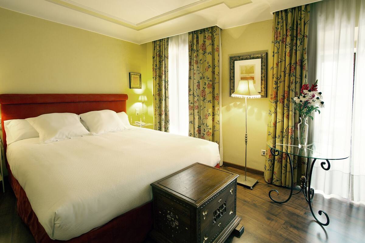 A neatly made bed with white linens, a wooden nightstand, and a glass table with a vase of flowers.