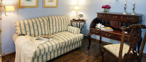 A room with a striped sofa, a wooden side table with a lamp, a wooden chair, and a dresser with a vase of flowers.