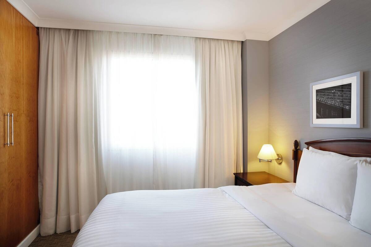 Una habitación de hotel con una cama grande, un armario de madera, una mesita de noche con una lámpara y un cuadro enmarcado en la pared.
