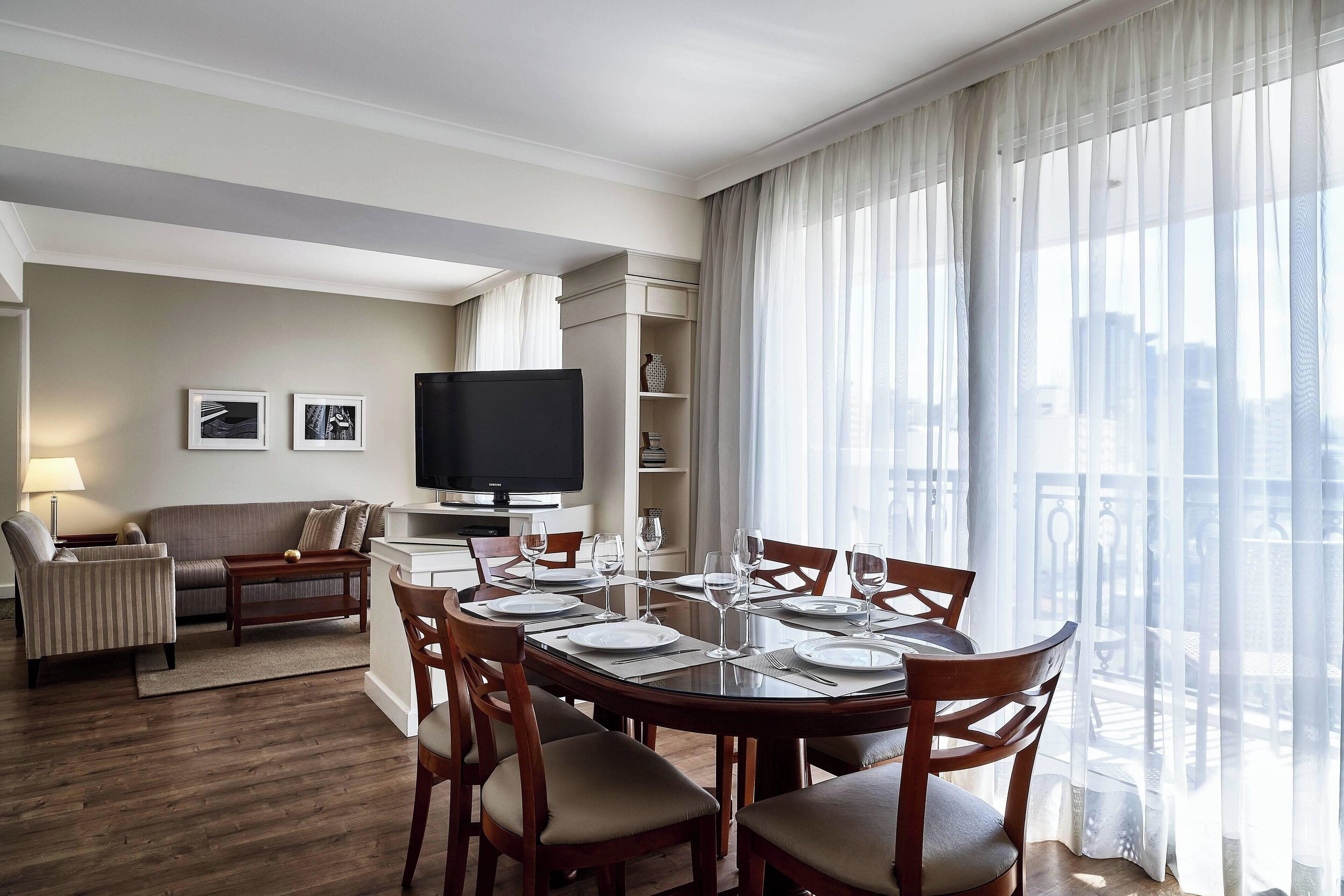 A modern hotel room with a dining area, a sofa, a TV, and a balcony with curtains.