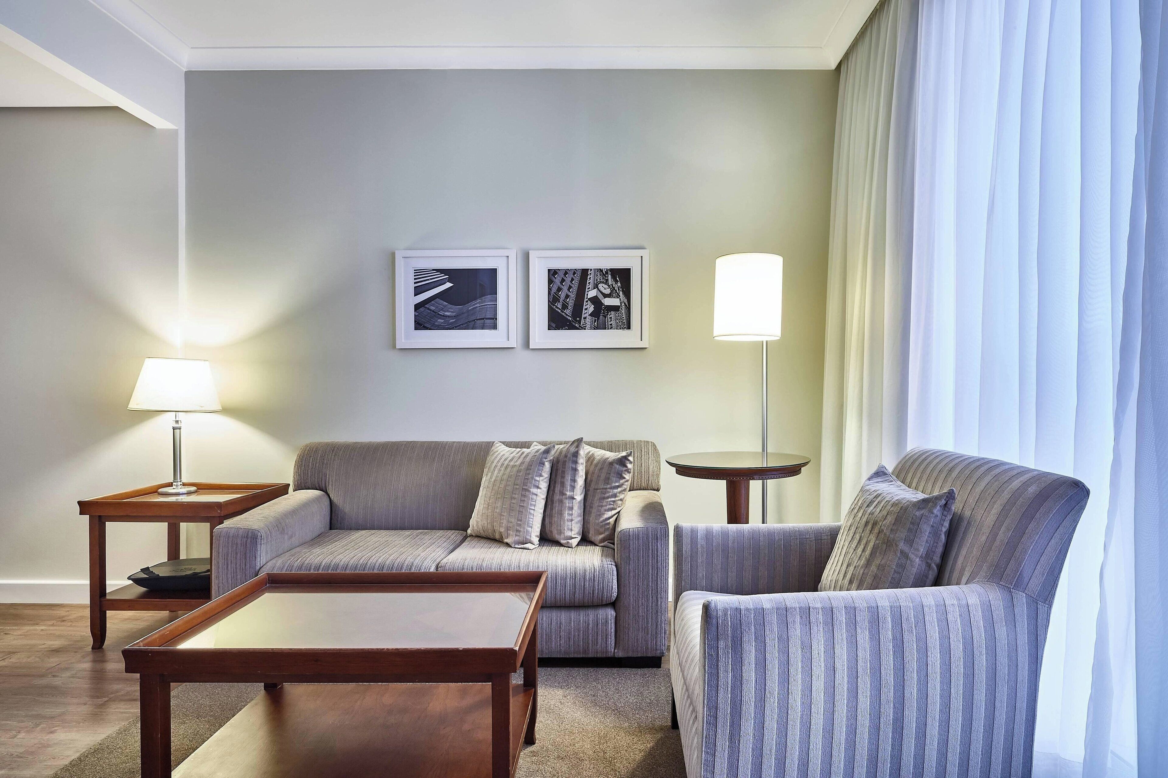 A modern living room with a sofa, armchair, coffee table, and two side tables. There are two framed pictures on the wall and a lamp on one of the side tables.