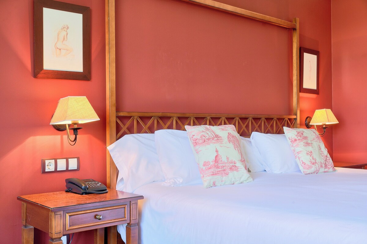 Habitación de hotel con cama, lámparas de noche, mesita de noche con teléfono y un cuadro en la pared.