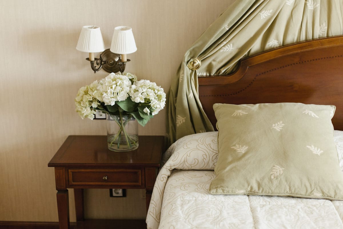 Una cama bien hecha con un cojín estampado y un florero con flores blancas sobre una mesita de noche de madera.