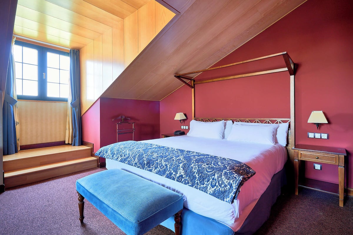 Un dormitorio con cama con dosel, un banco, una mesita de noche y una ventana con cortinas.