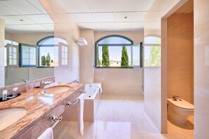 Un baño moderno con un espejo grande, dos lavamanos, una bañera y un inodoro. Ventanas en arco ofrecen vistas a árboles y un paisaje lejano.