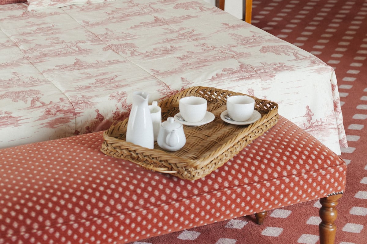 Una cama bien hecha con colcha roja y blanca de lunares, cabecera de madera y mesita de noche con una lámpara y una pequeña planta.