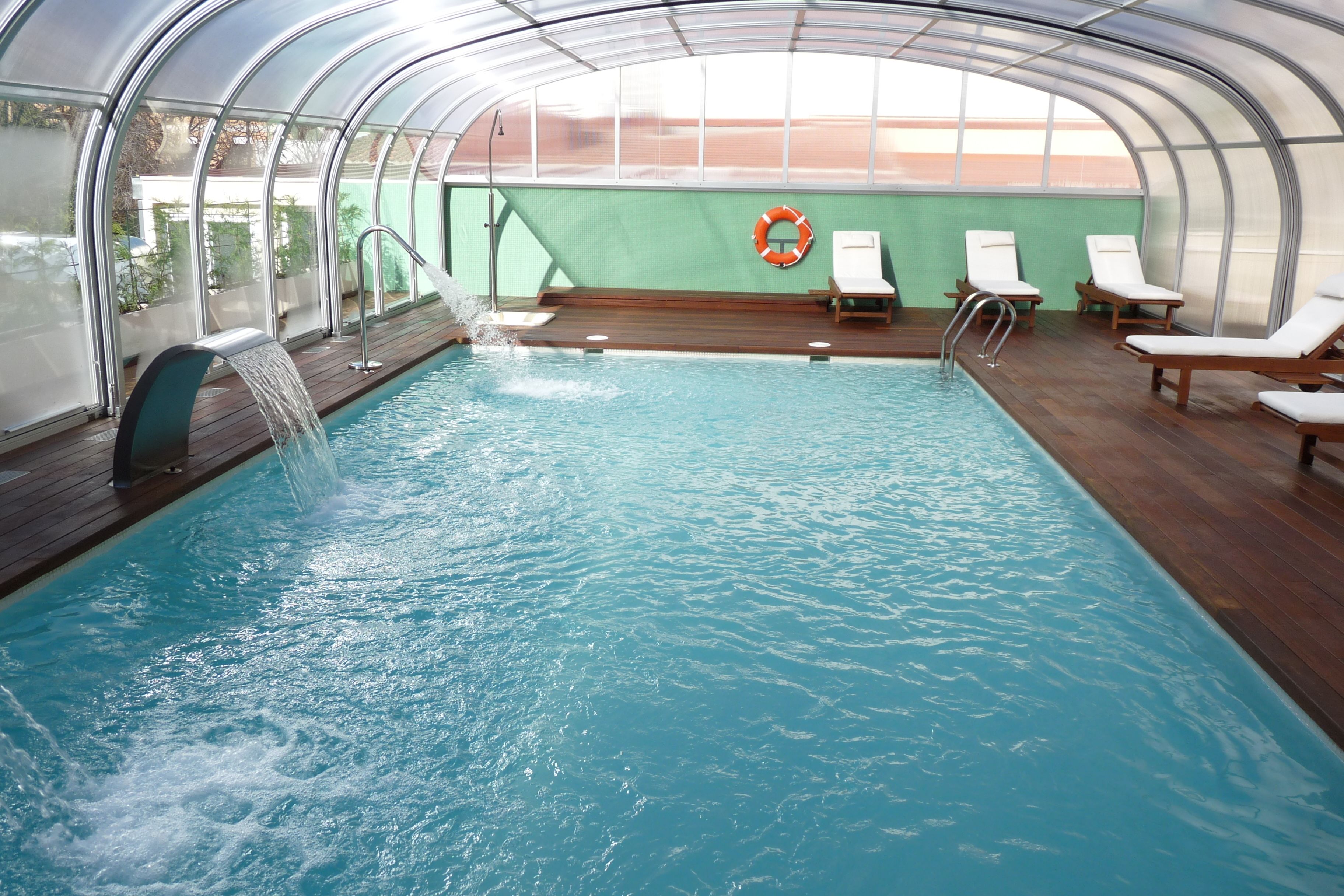 Una piscina interior con agua cristalina, terraza de madera y sillones de descanso.
