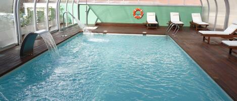 Una piscina interior con agua cristalina, terraza de madera y sillones de descanso.