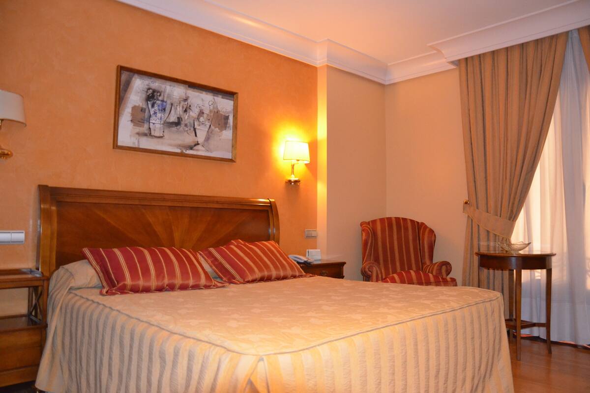 A hotel room with a large bed, a wooden headboard, a framed picture on the wall, a standing lamp, a striped armchair, and a small table with a bowl.