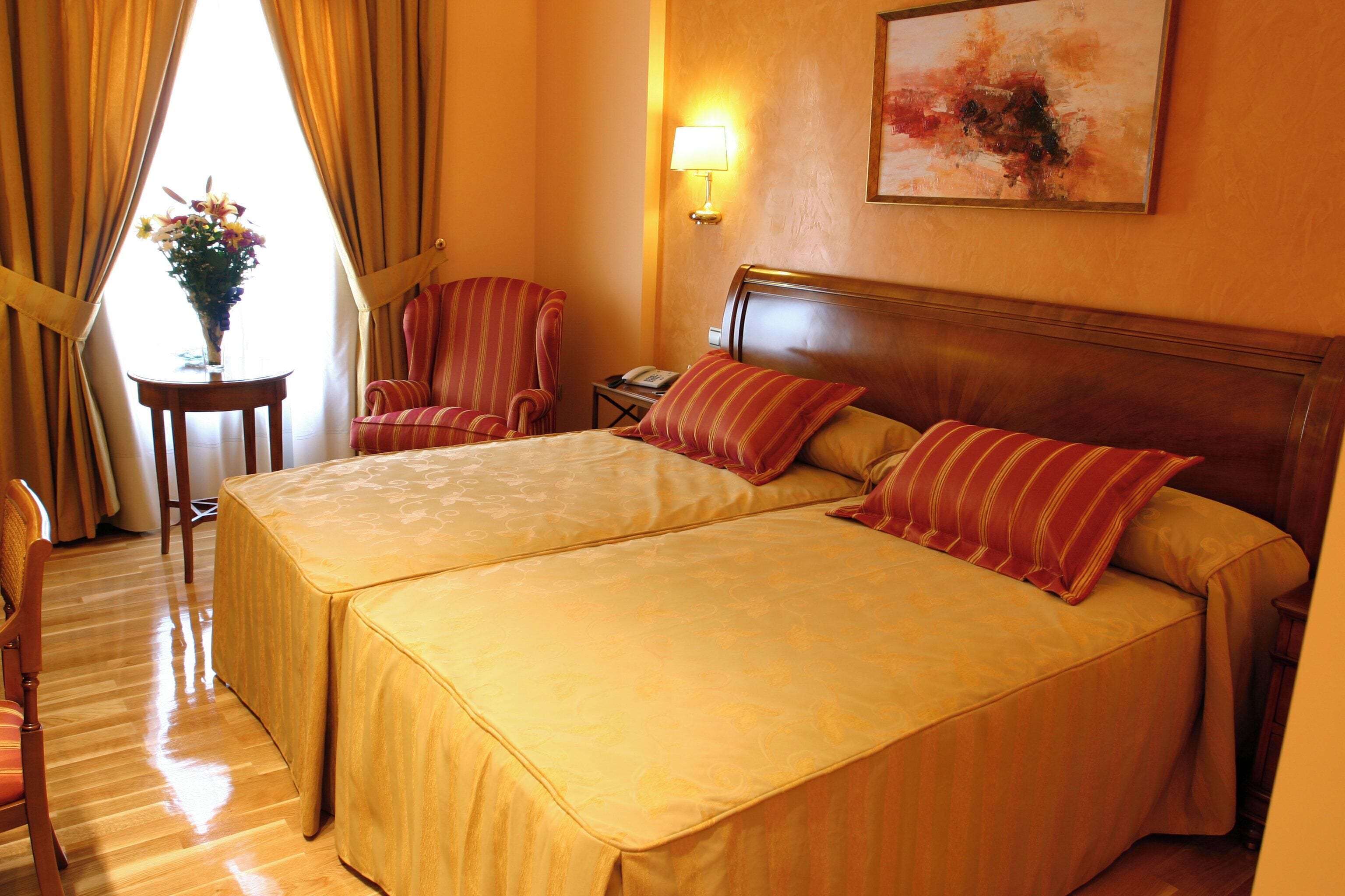 A hotel room with two beds, a sitting area with a chair, a small table with a vase of flowers, and a lamp on the wall.