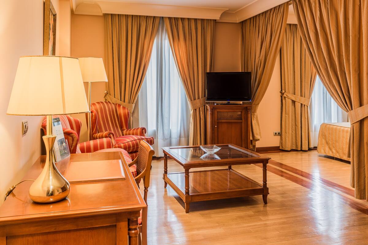 A hotel room with a wooden coffee table, a sofa, a TV on a stand, and curtains.