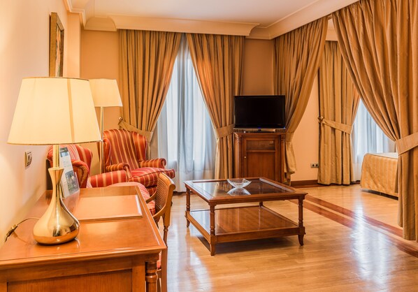 Una habitación de hotel con una mesa de centro de madera, un sofá, un televisor sobre un mueble y cortinas.
