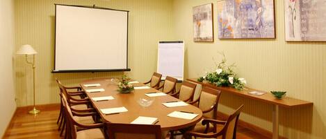 A conference room with a long table, chairs, a projector screen, a whiteboard, and framed artwork on the walls.