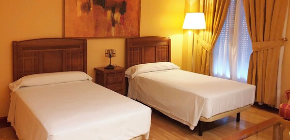Habitación de hotel con dos camas individuales, una mesita de noche con lámpara, un cuadro en la pared y una ventana con cortinas.