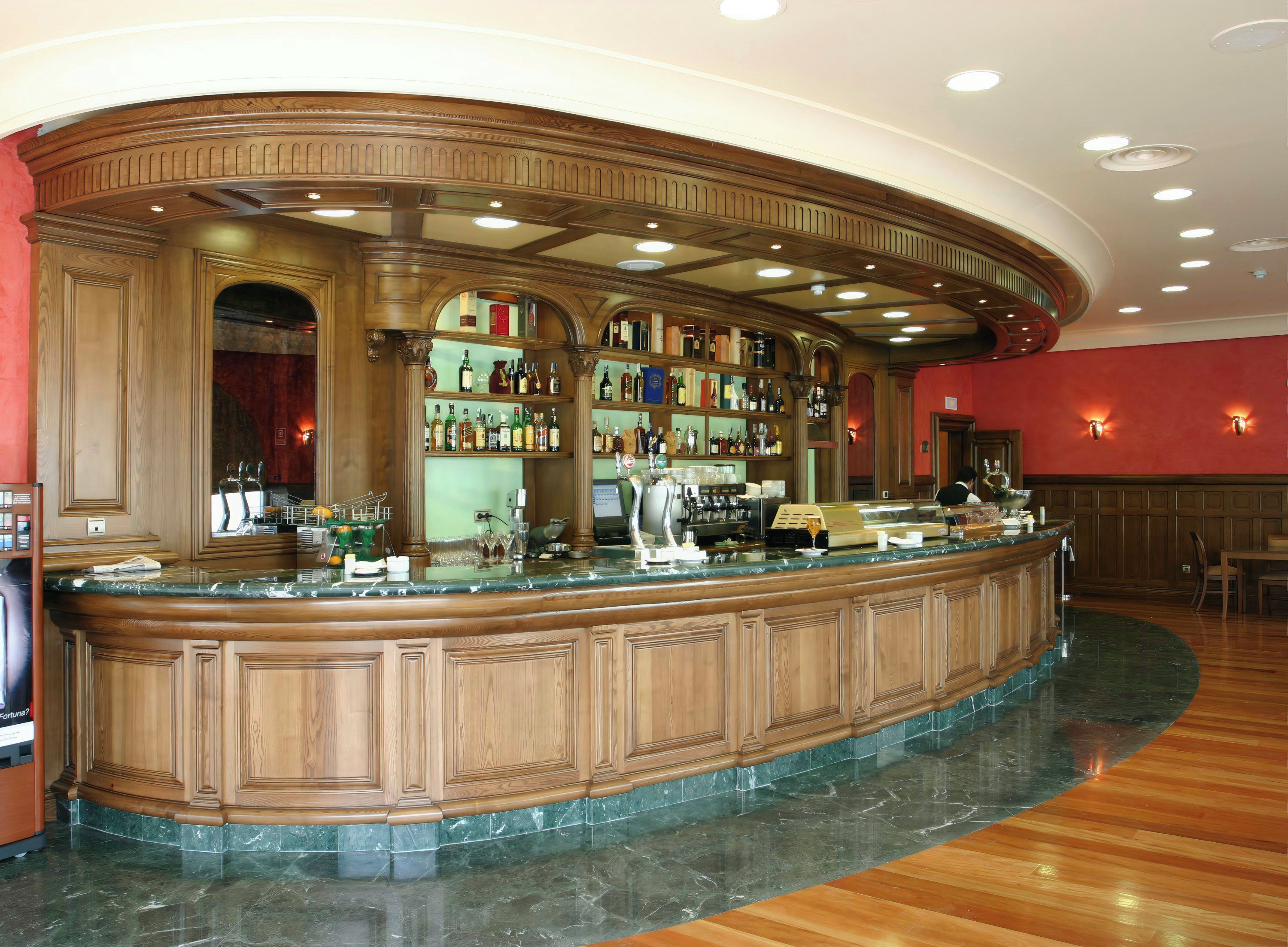 Zona de bar con mostrador de madera curvo, estantes con botellas y una caja registradora.