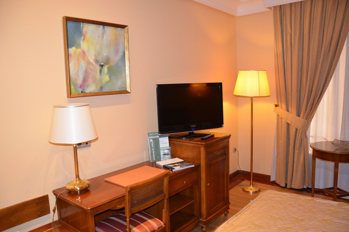 A hotel room with a television, a desk with a lamp, a chair, a small table, and a decorative painting on the wall.