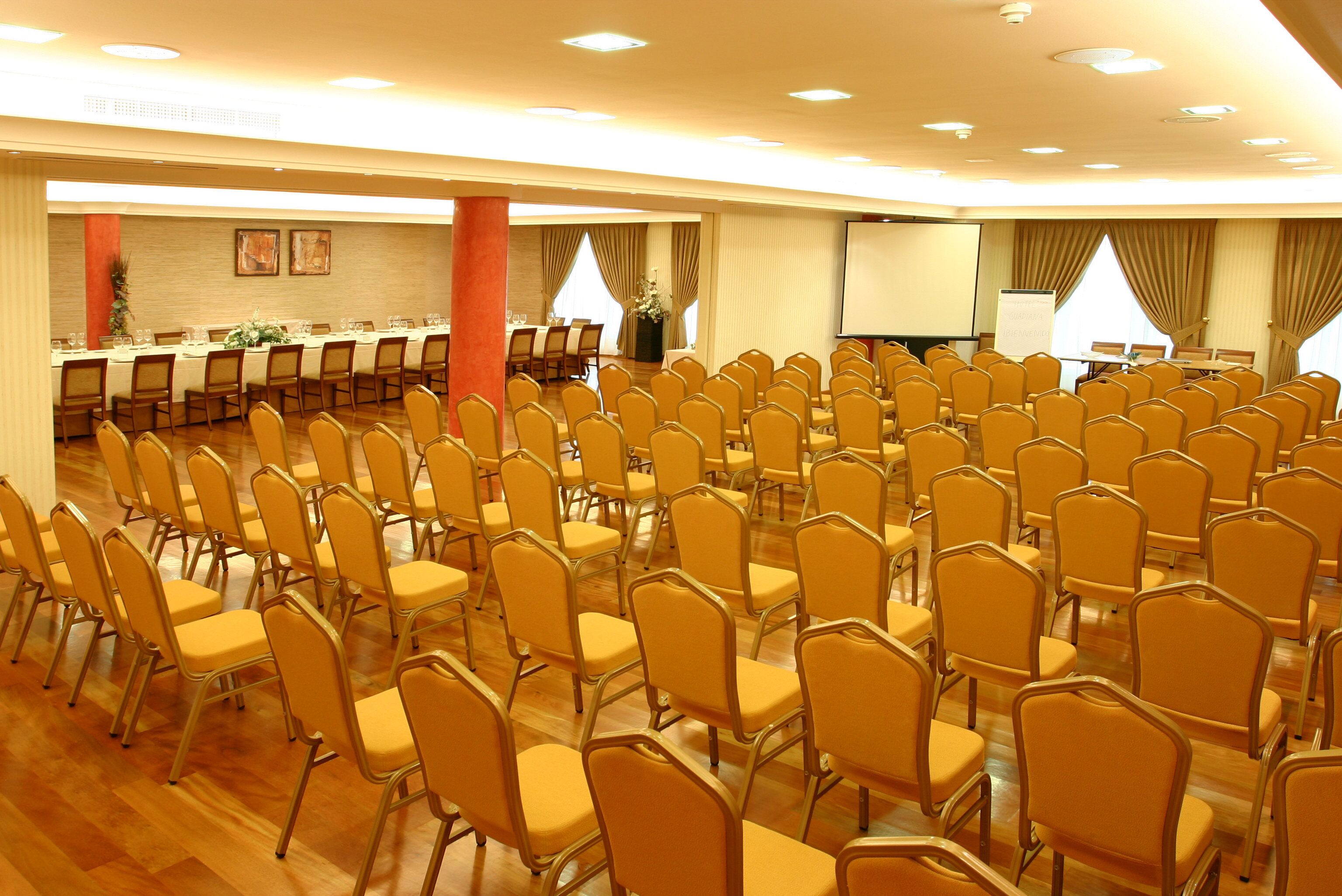 Un salón de conferencias con filas de sillas amarillas, una mesa larga con sillas, una pantalla de proyector y una pizarra.