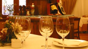 Una zona de bar con estantes llenos de botellas de licor y vasos, una mesa con copas de vino y platos vacíos, y un ventanal con cortinas.
