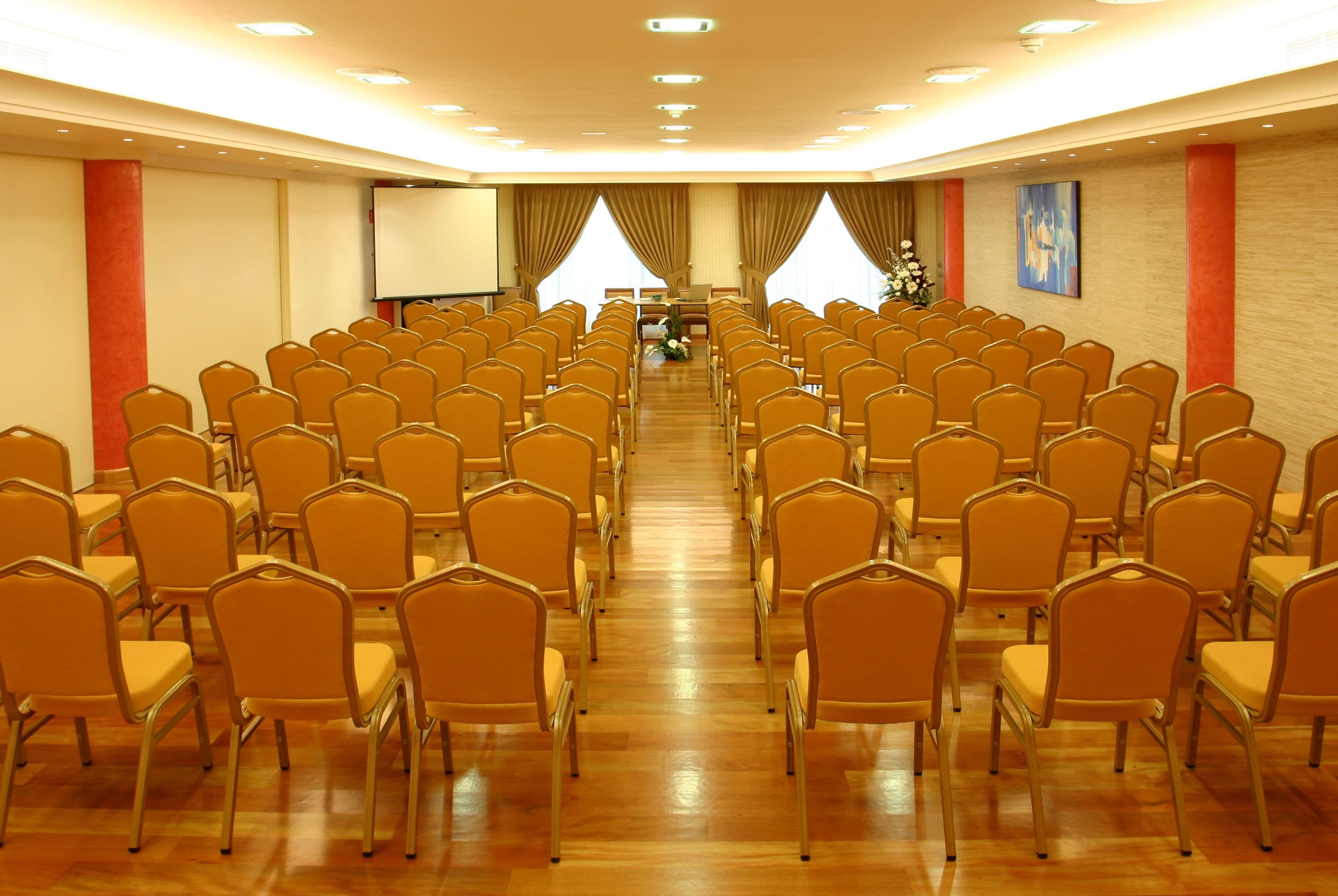 Un salón de conferencias con filas de sillas orientadas hacia un escenario, una pantalla de proyector y un arreglo floral.
