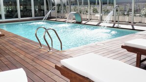 Una piscina con dos chorros de agua, terraza de madera y sillones de descanso.