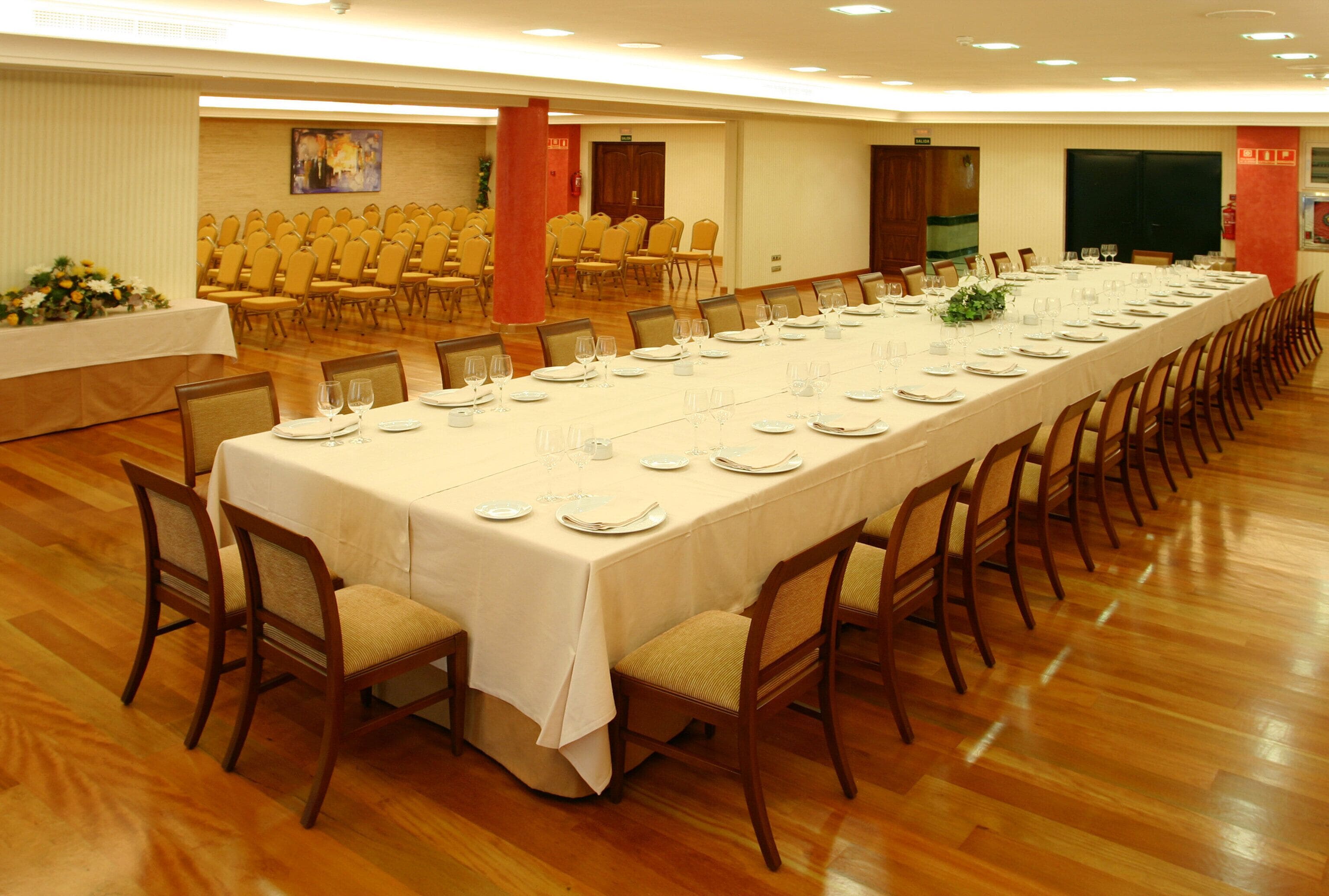 A long banquet table set for a formal event, surrounded by wooden chairs, in a spacious room with wooden flooring and a high ceiling.