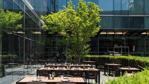 An outdoor dining area with tables and chairs, a glass building in the background, and a tree in the center.