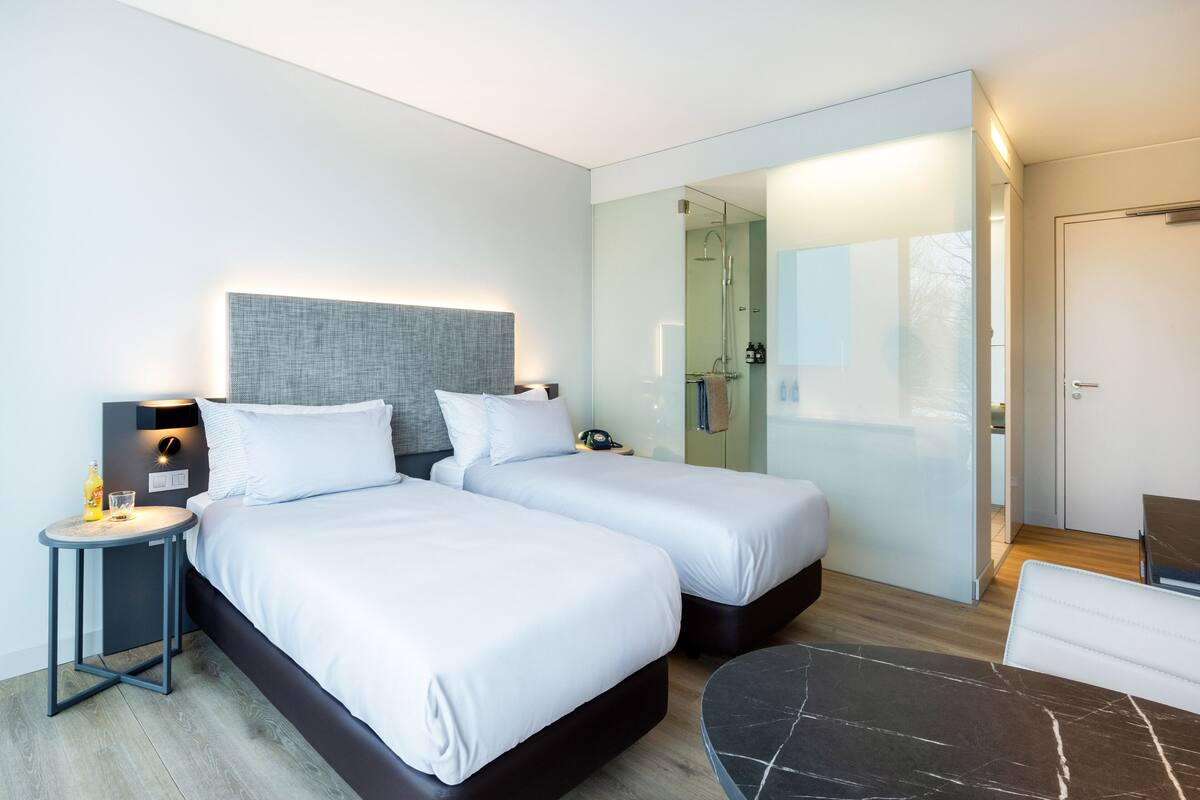 Une chambre d’hôtel moderne avec deux lits, une petite table, un coin salon et une douche à parois de verre.