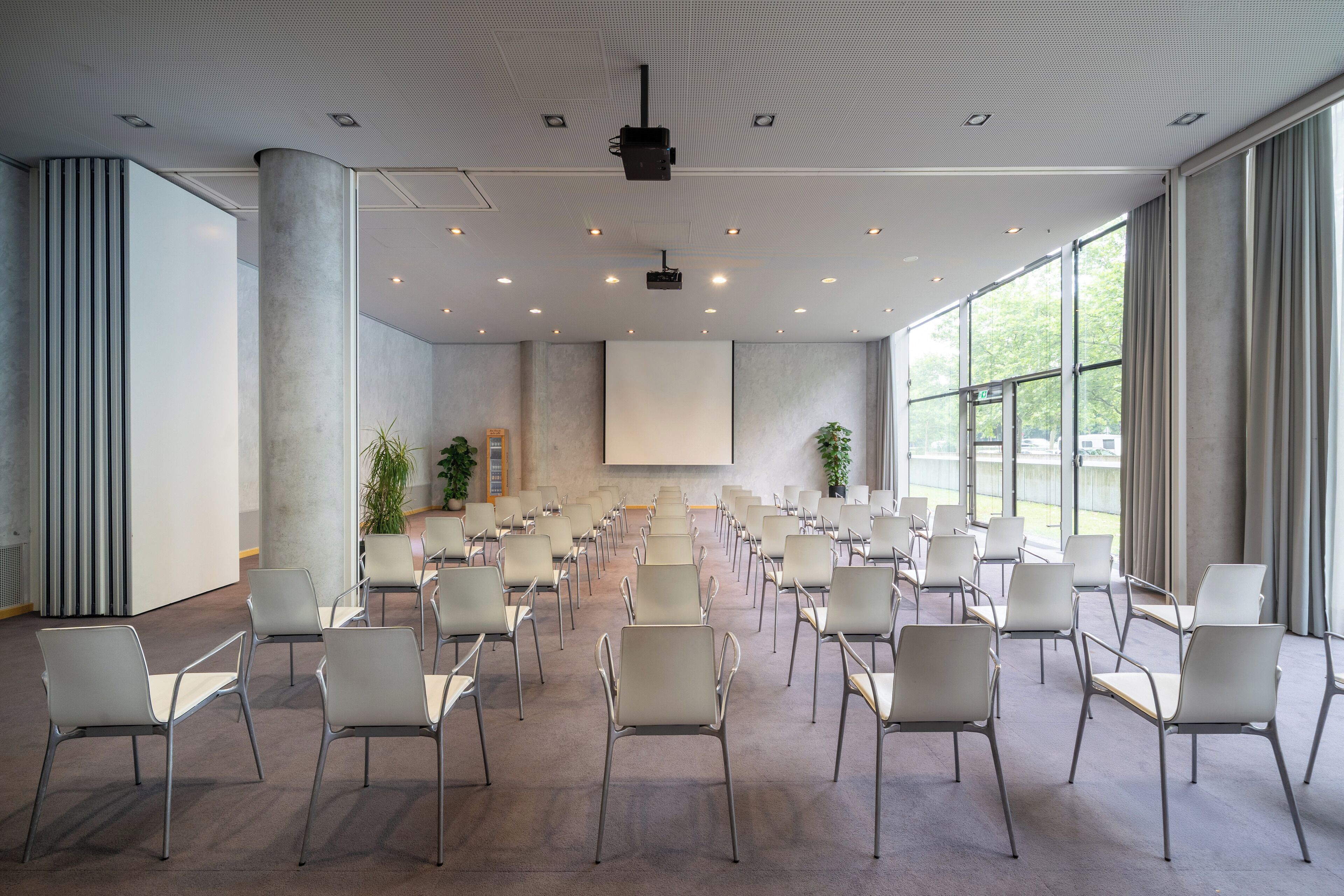 Un salón de conferencias con filas de sillas, una pantalla de proyector y grandes ventanales.