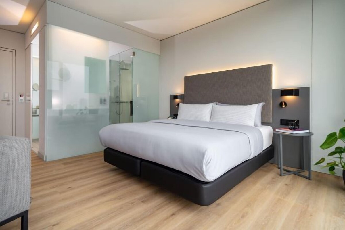 A modern bedroom with a bed, bedside tables, a glass shower enclosure, and a potted plant.