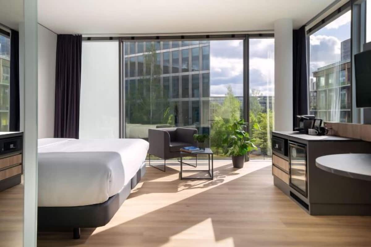 A modern hotel room with a large bed, a small desk, a chair, a potted plant, and a view of the city.