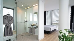 A modern hotel room with a glass shower enclosure, a bedside table with toiletries, a bed with a white mattress, and a large window with blinds.