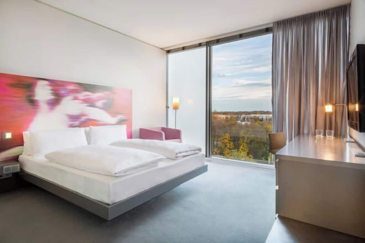 Une chambre d’hôtel moderne équipée d’un lit, d’une table de chevet et d’une grande fenêtre donnant sur la ville.