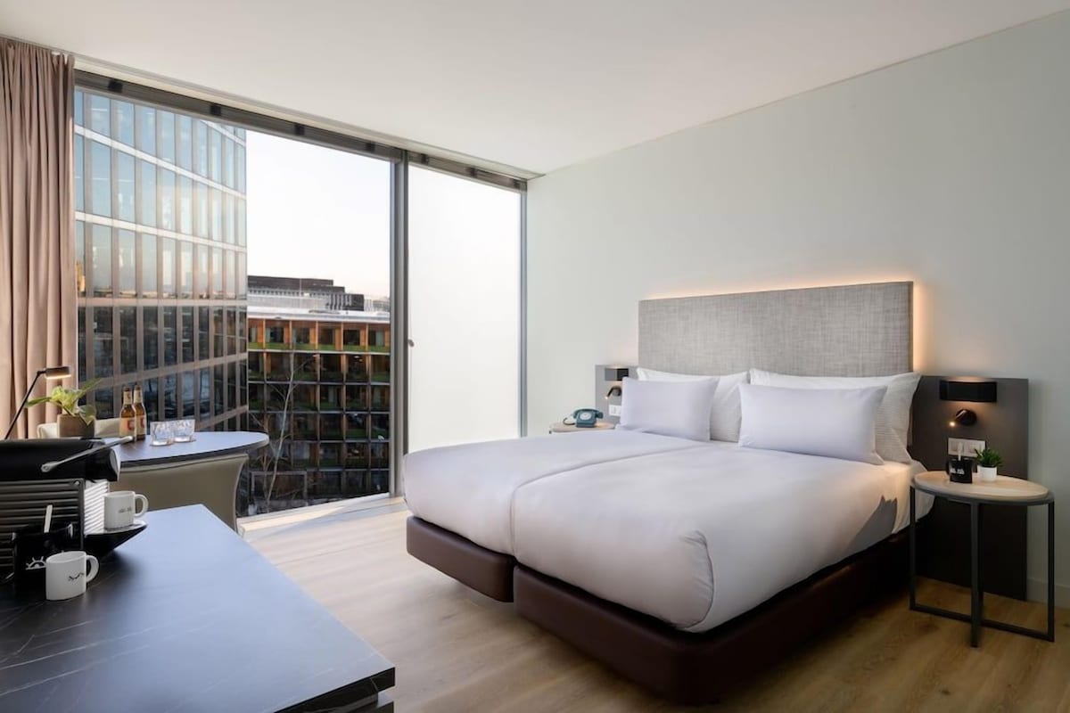 A modern hotel room with a large bed, a desk with a chair, a small table, and a view through a large window.
