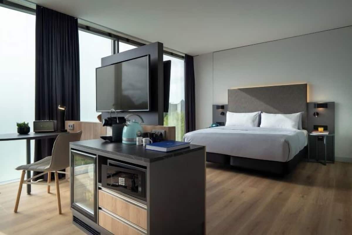 A modern hotel room with a bed, a desk with a TV, a kitchenette, and a view of the outdoors.