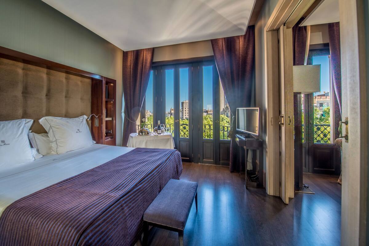A hotel room with a large bed, a bench, a TV, and a balcony with a view of the city.