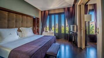 Habitación de hotel con una cama grande, un banco, un televisor y un balcón con vistas a la ciudad.
