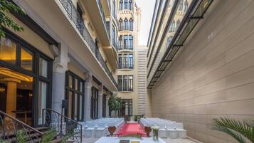 Un estrecho patio con una alfombra roja que conduce a una mesa blanca, rodeada de sillas blancas, y flanqueado por edificios con balcones y ventanas amplias.