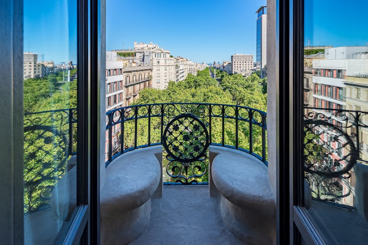A balcony with a wrought iron railing, a stone bench, and a view of a cityscape with buildings and trees.