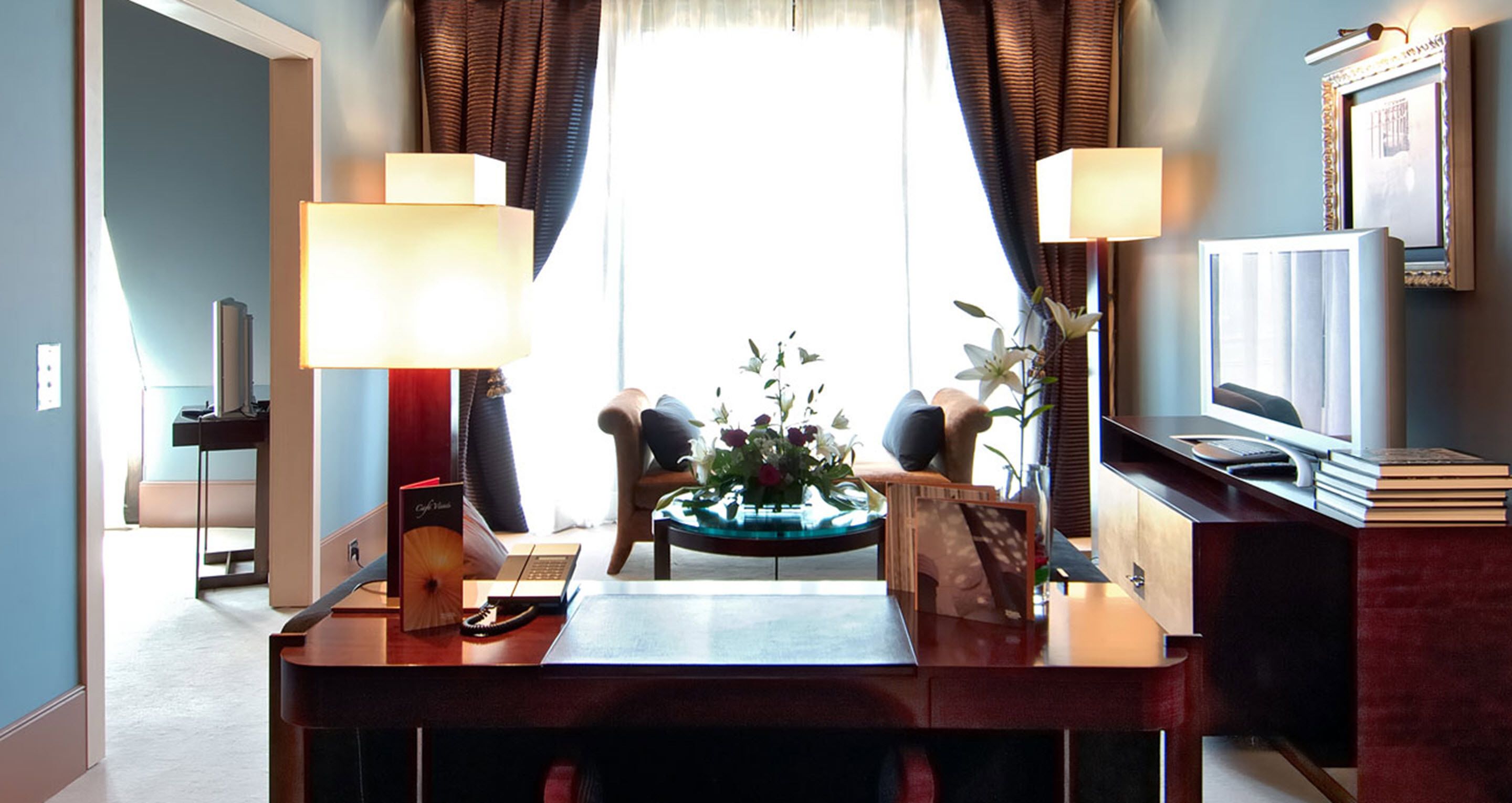 Habitación de hotel con escritorio, dos lámparas, televisión, estantería y ventana con cortinas.