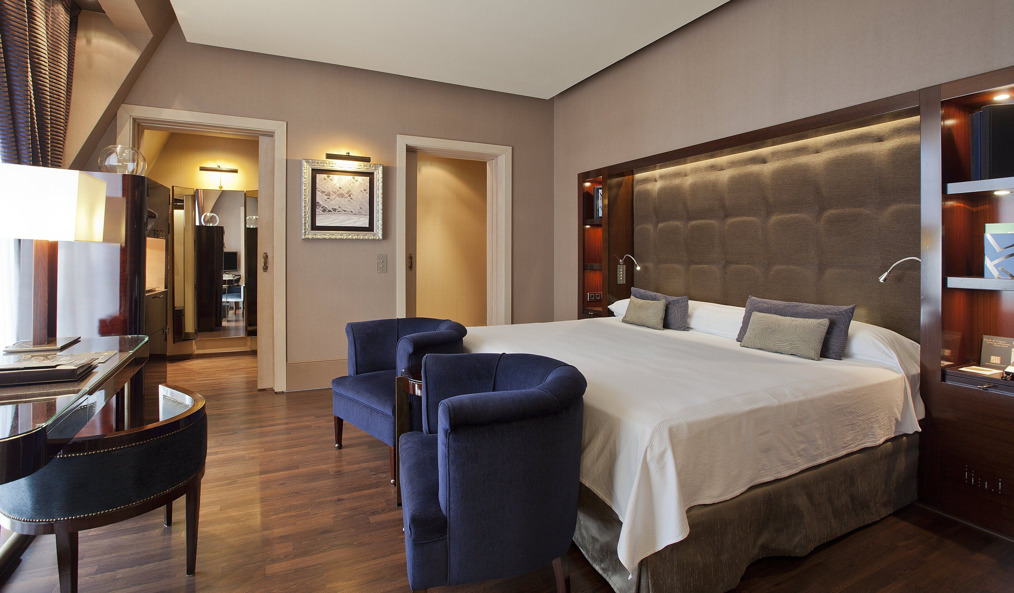 A hotel room with a large bed, two armchairs, a desk with a chair, and a mirror.