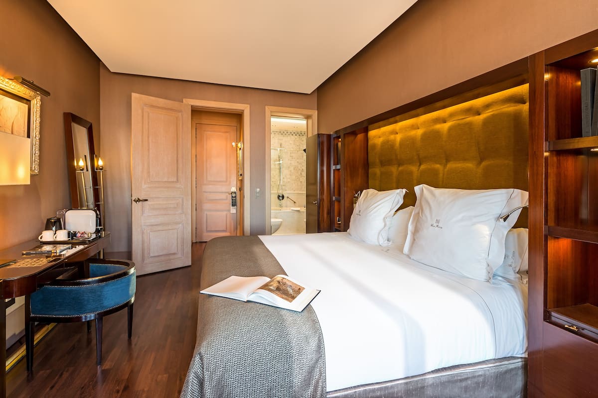 A hotel room with a large bed, a desk with a chair, a mirror, and a bathroom visible through an open door.