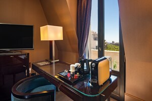 A hotel room with a desk, a lamp, a coffee maker, and a view of greenery outside.