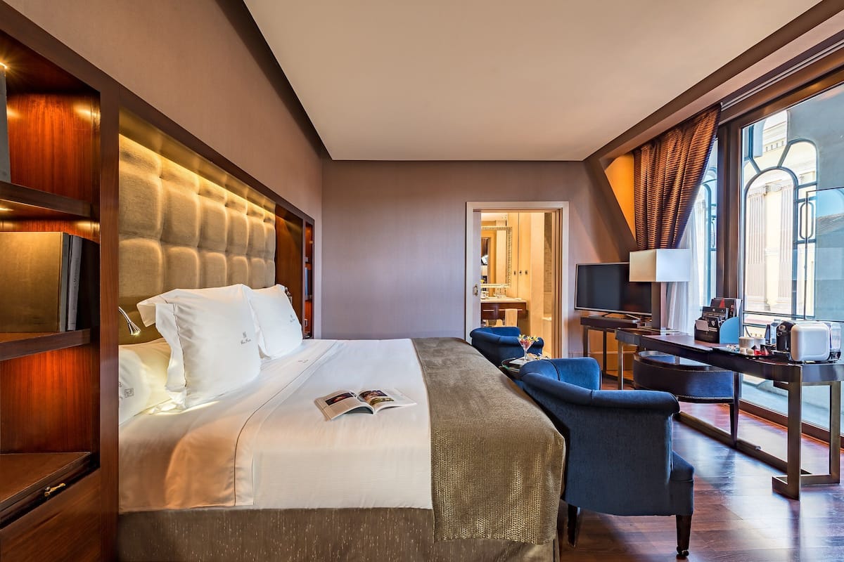 A modern hotel room with a large bed, a desk, and a view of the city through large windows.