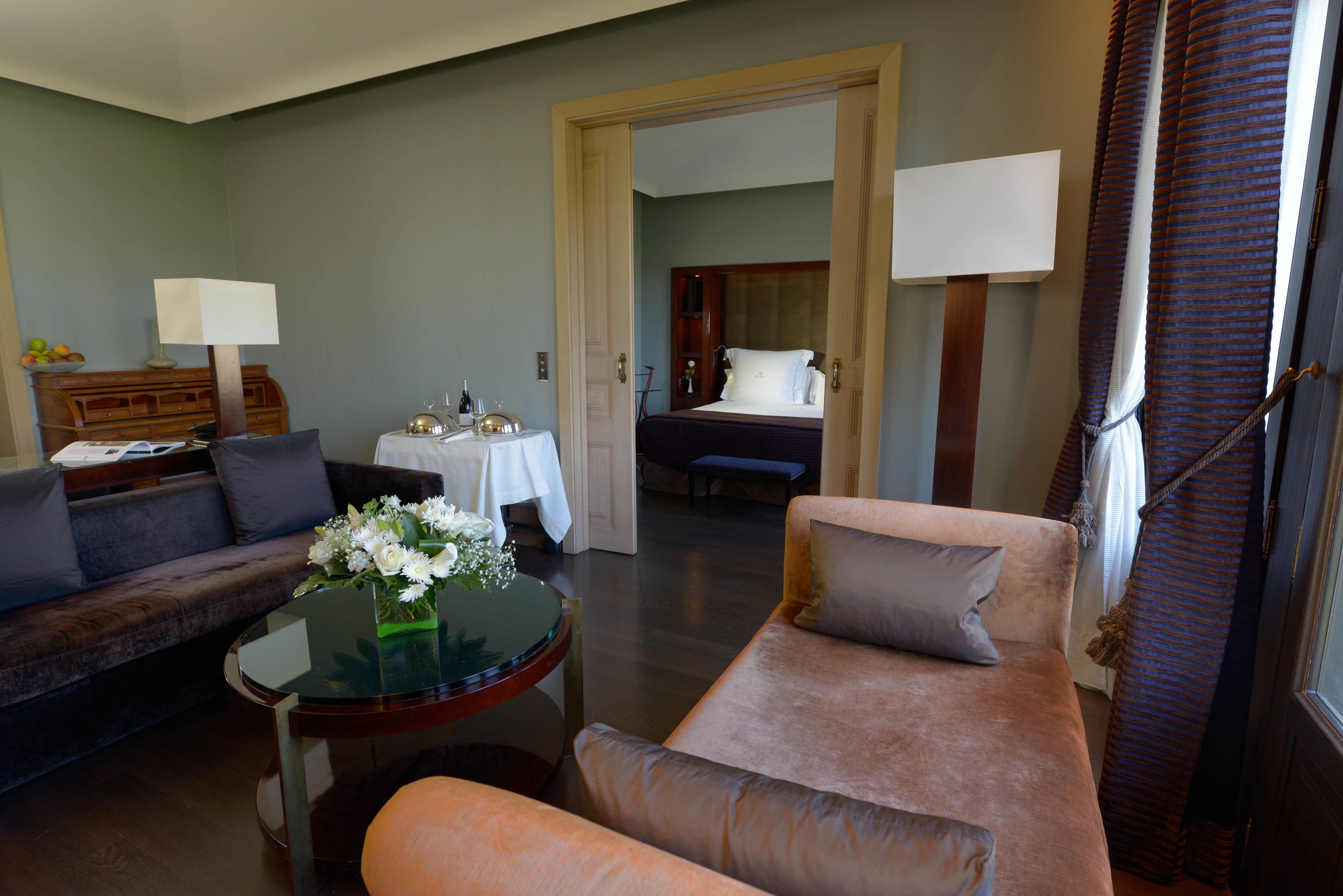 A hotel room with a living area featuring a sofa, a coffee table with flowers, a dining table with a lamp, and a bed in the background.