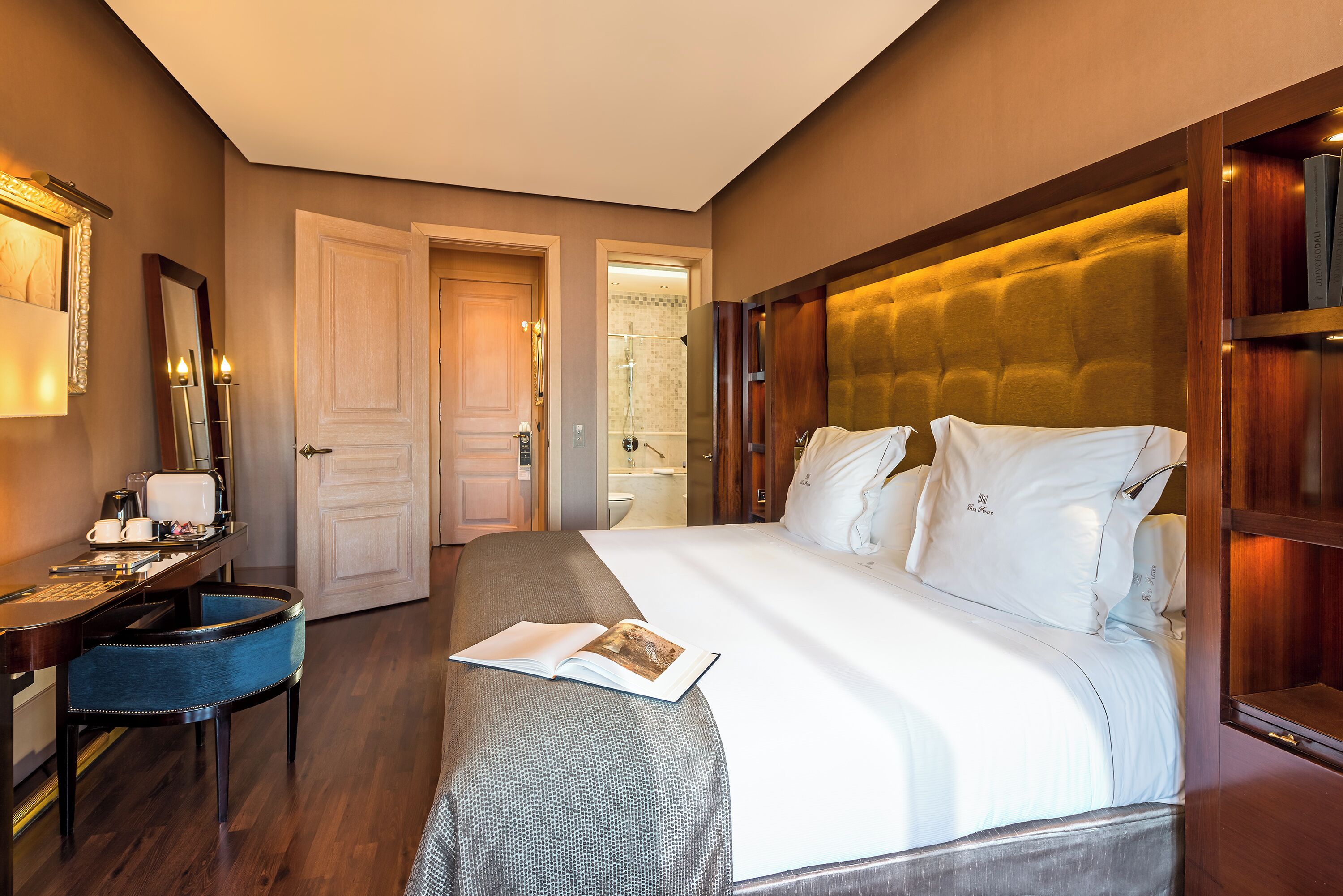 Een hotelkamer met een groot bed, een bureau met stoel, een spiegel en een deur naar de badkamer.