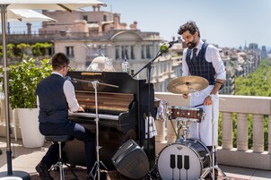 A musician playing a piano and drums on a balcony with a city view.