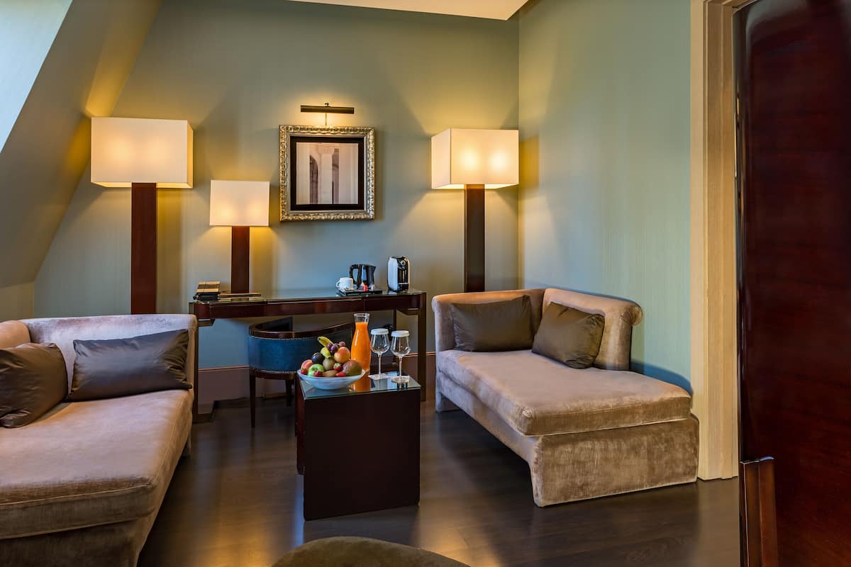 A modern hotel room with two sofas, a coffee table with fruits and glasses, and two wall-mounted lamps.